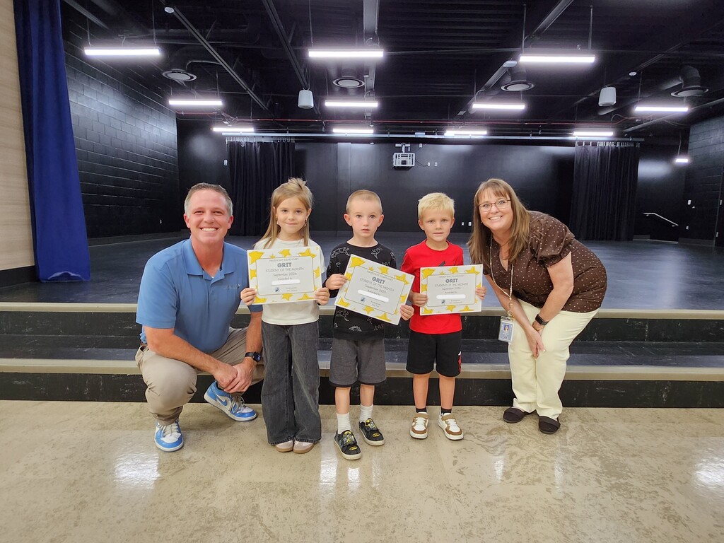 Mr. Skeen crouching and a Mrs. Widdison standing alongside three young students holding "GRIT - Student of the Month - September 2024" certificates on stage.