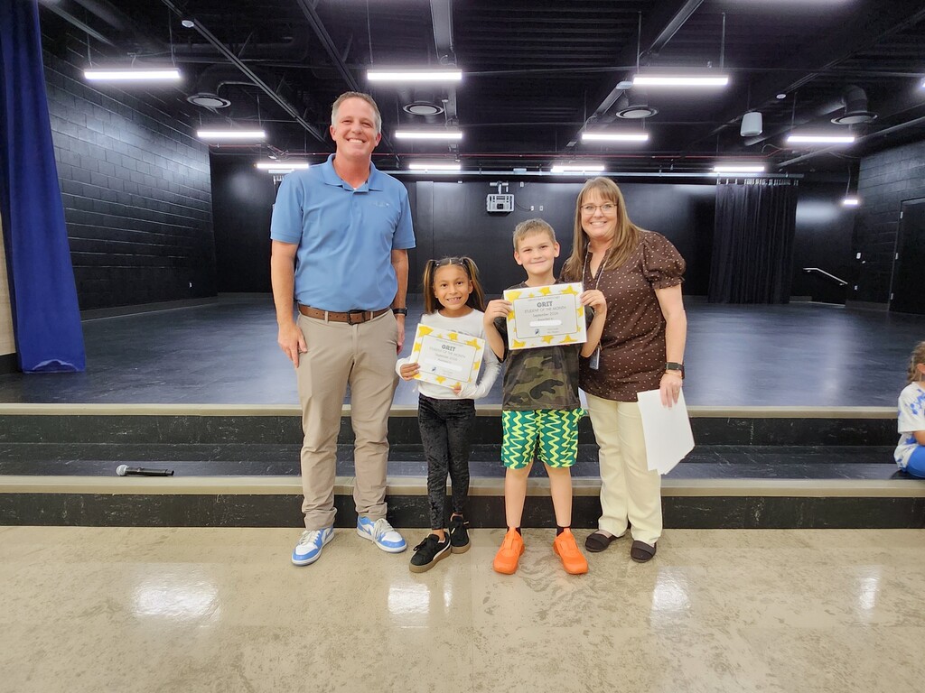 Mr. Skeen and a Mrs. Widdison standing alongside two third grade students holding "GRIT - Student of the Month - September 2024" certificates on stage.