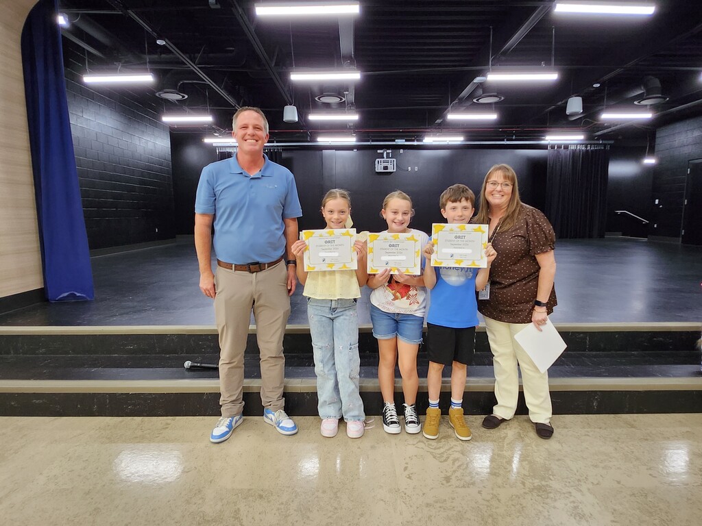 Mr. Skeen and a Mrs. Widdison standing alongside three fifth grade students holding "GRIT - Student of the Month - September 2024" certificates on stage.