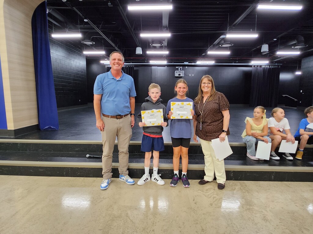 Mr. Skeen and a Mrs. Widdison standing alongside three sixth grade students holding "GRIT - Student of the Month - September 2024" certificates on stage.