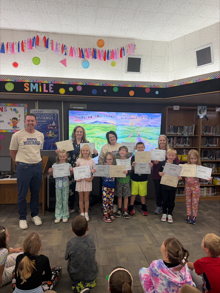Mr. Karlinsey and two staff members stand with a group of elementary students in the school library. The students hold certificates recognizing their summer reading and math achievements while classmates sit on the floor watching. Colorful decorations and a large “SMILE” banner are visible in the background.