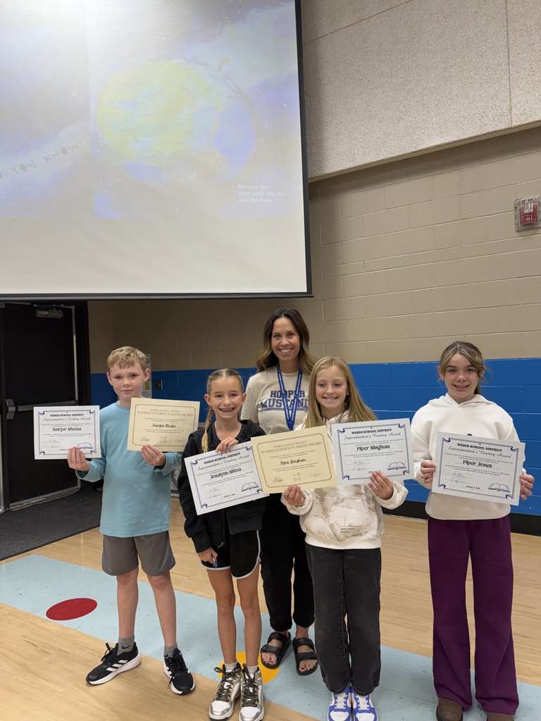 A staff member stands with four elementary students in a school gym. Each student holds a certificate recognizing their summer reading achievement, smiling for the photo.