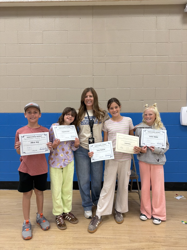 A staff member stands with four elementary students in a school gym. Each student holds a certificate recognizing their summer reading achievement, smiling for the photo.