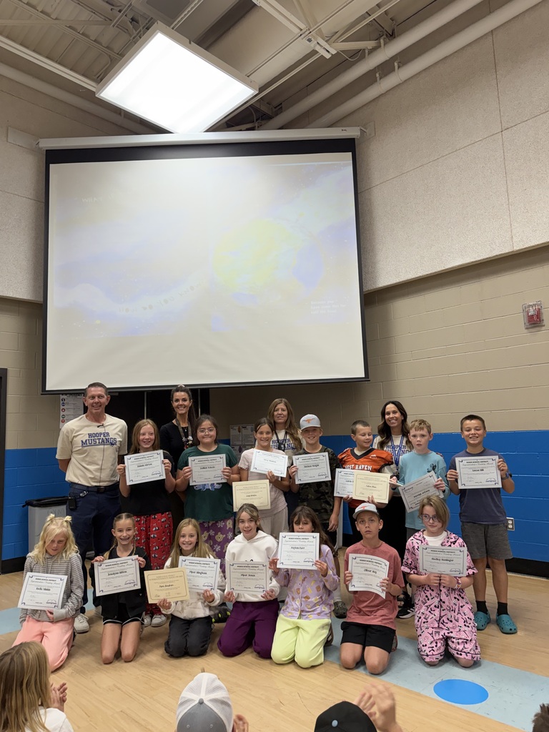 Mr. Karlinsey, four staff members, and a group of elementary students pose in the school gym while holding certificates recognizing their summer reading and math achievements. Other students sit in the foreground watching and applauding.