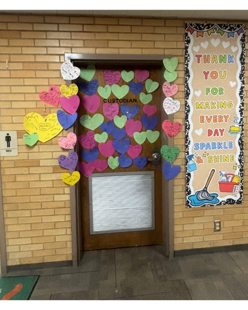 A wooden hallway door with a silver kickplate, labeled "CUSTODIAN" at the top. The door and surrounding brick wall are decorated with dozens of colorful, heart-shaped paper cutouts in pink, green, yellow, blue, and purple, each filled with handwritten thank-you notes. To the right of the door hangs a bulletin board framed with colorful pennants and border trim. The board features white and yellow lettering that reads: "THANK YOU FOR MAKING EVERY DAY SPARKLE & SHINE," decorated with illustrations of a broom, mop bucket, spray bottles, and soap bubbles.