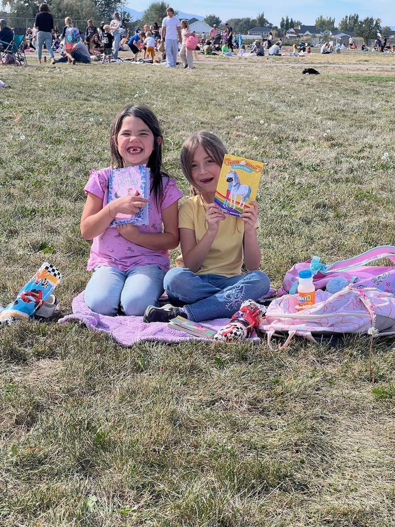 two girl students holding up books sitting on a blanket on the grass smiling 