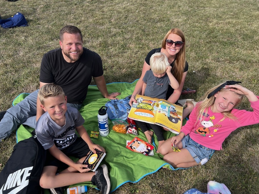 family reading books with their children on a blanket on the grass all are looking at the camera smiling 
