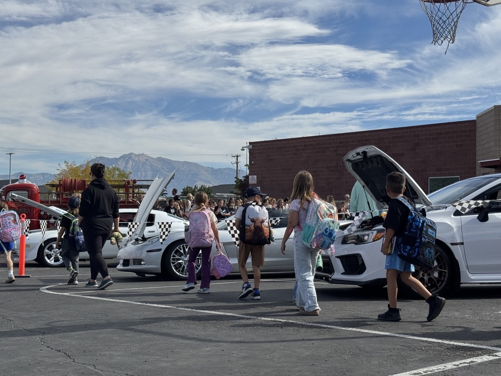 old cars in a line and a line of students walking next to them, only the back of the students heads are shown