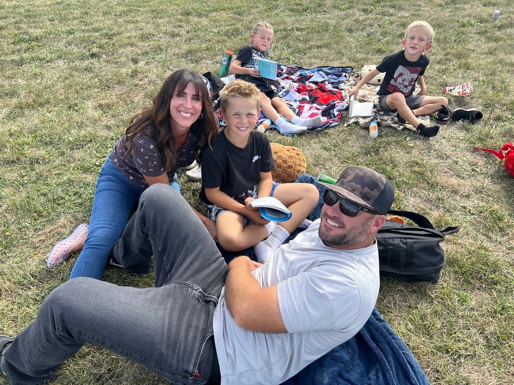 family reading books with their children on a blanket on the grass all are looking at the camera smiling 