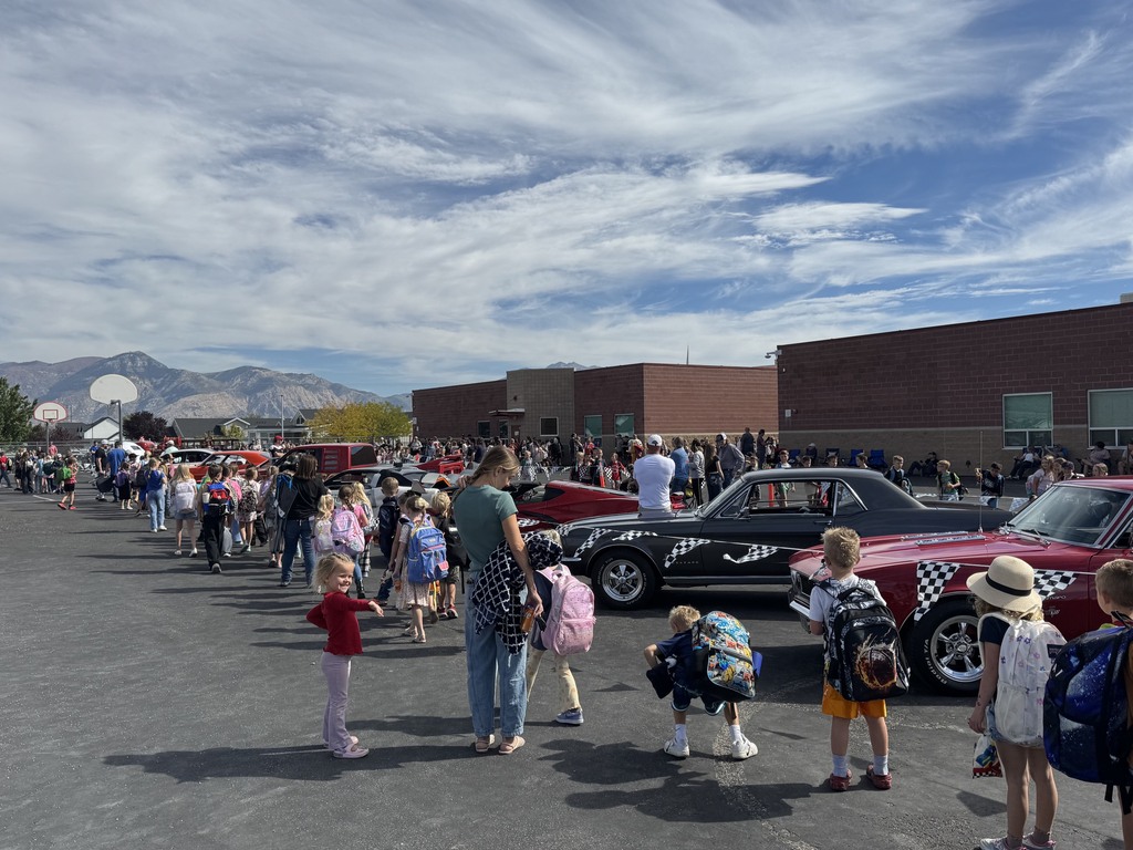 old cars in a line and a line of students walking next to them, only the back of the students heads are shown