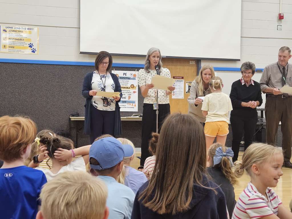 A wider audience perspective looking over seated students. A line of administrators stands on the gym floor, and a young student walks across the floor towards them to receive a certificate