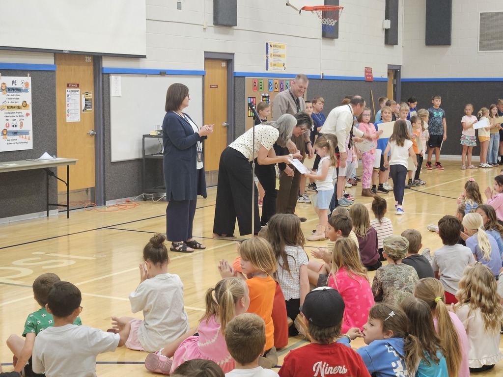 A group of administrators stands center stage, clapping. Four students stand in a line to the right, proudly holding up their newly received awards