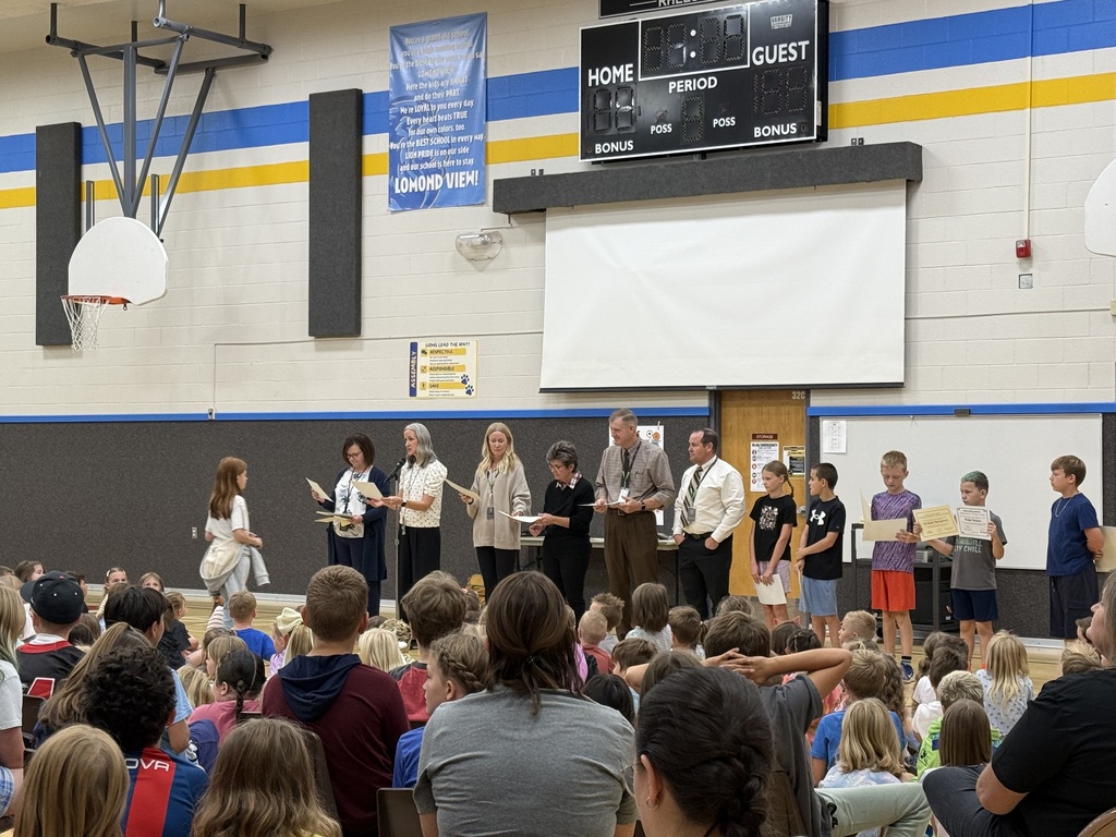 A wider audience perspective looking over seated students. A line of administrators stands on the gym floor, and a young student walks across the floor towards them to receive a certificate