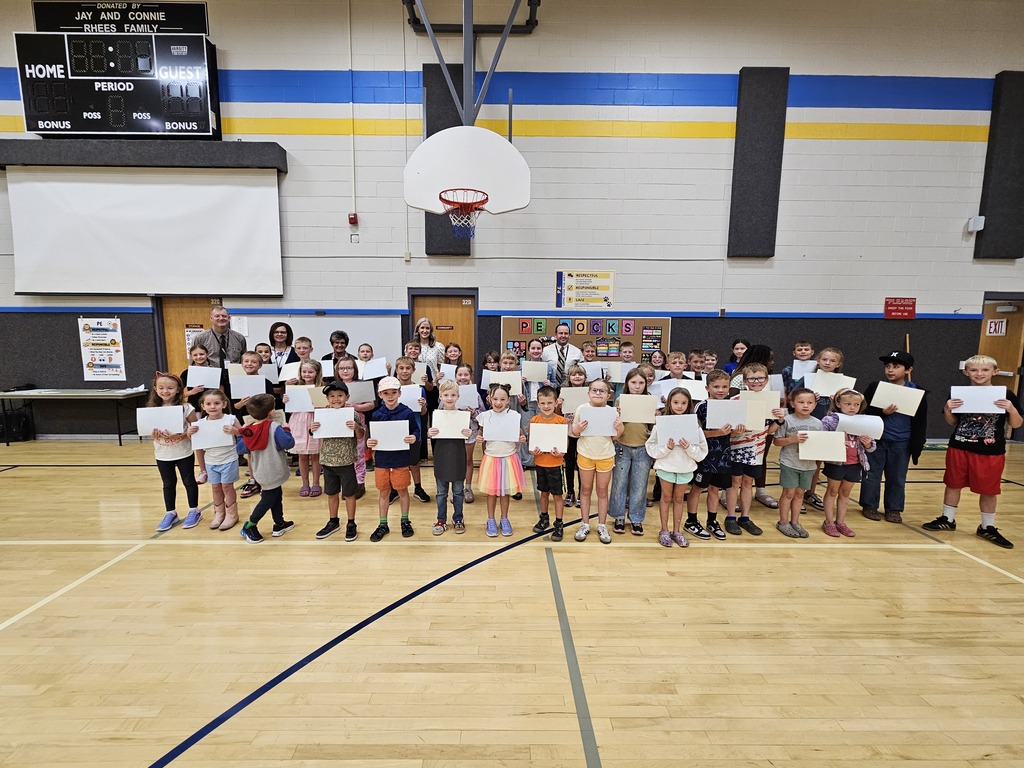 A wide-angle landscape photograph of dozens of elementary school students standing in three rows across a wooden gym floor, proudly holding up white certificates for a group photo. Several adults, likely teachers or administrators, stand behind them. The school logo "Lomond View" is visible on the wall behind them