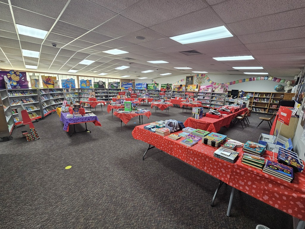A wide view of the Lomond View Elementary Library transformed for the Scholastic Book Fair, featuring tables lined with red tablecloths, display cases, and stacks of colorful new books ready for students to browse.