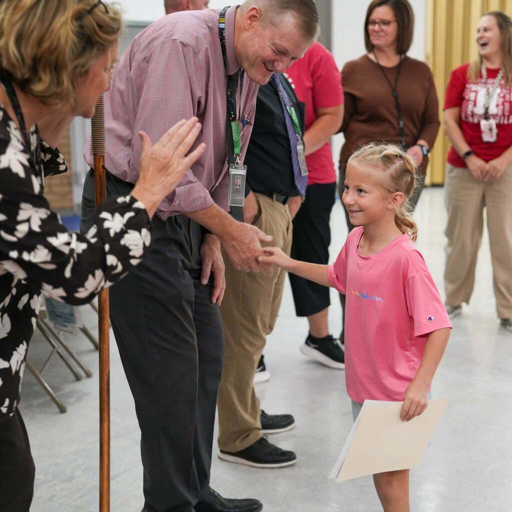 Image 7: A smiling young student wearing a pink shirt receiving a certificate, shaking hands with a school administrator, and being greeted by school staff.
