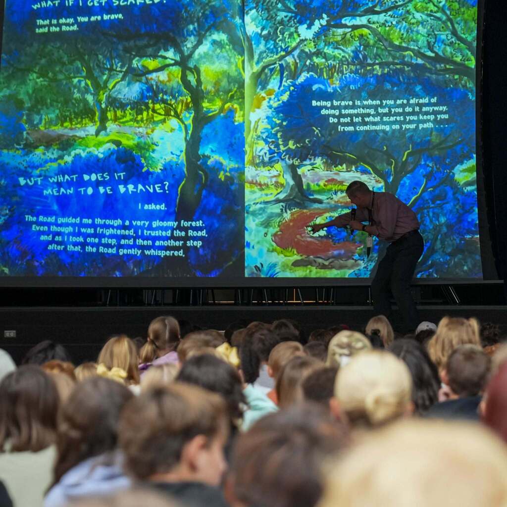 Image 4: A staff member pointing to a large projection screen displaying pages from a storybook about courage during an assembly in front of a seated audience of students.