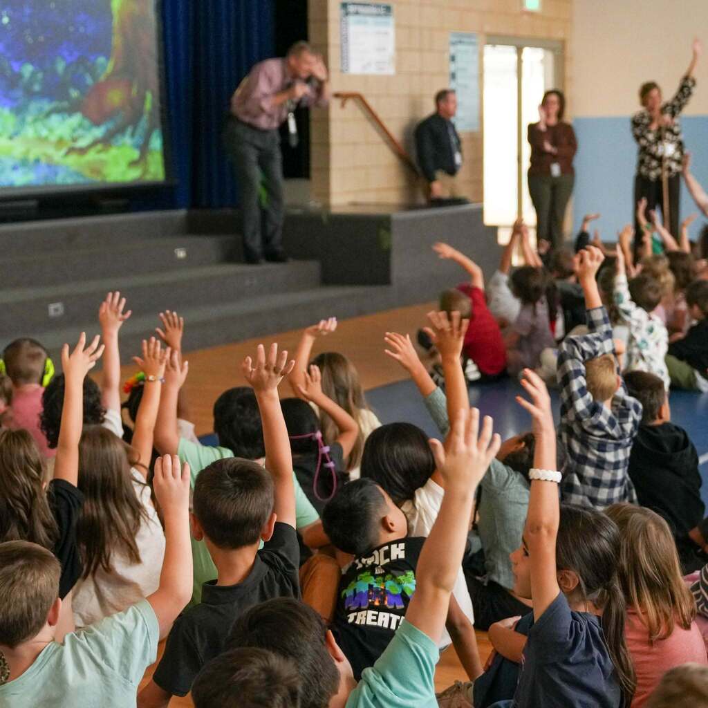 Image 5: Elementary students sitting on the floor with their hands raised high in the air to answer a question during an assembly with school administrators on stage.
