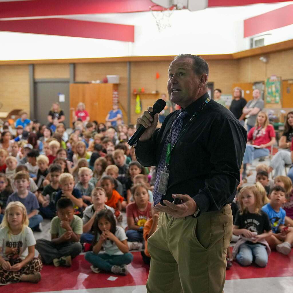 Image 6: A school administrator speaking into a microphone and gesturing with his hand while addressing a gym full of students seated on the floor.