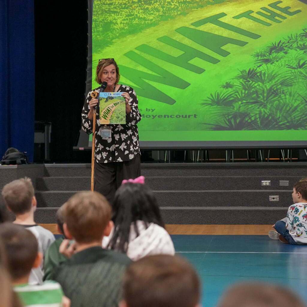 Image 2: An educator on stage holding up the picture book What the Road Said by Cleo Wade while speaking into a microphone in front of a large screen showing the book's cover artwork to students.