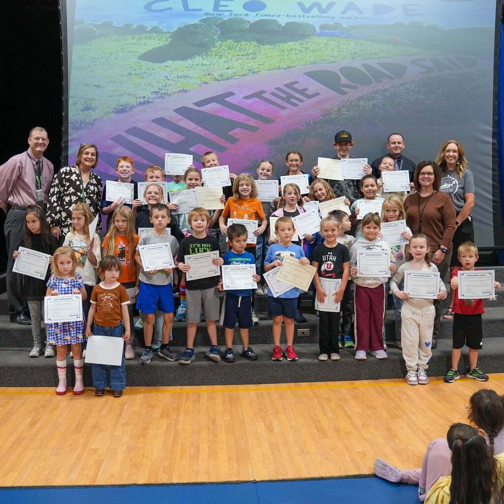 Image 9: A large group of elementary students and school staff posing together on stage, holding up their academic award certificates in front of a projection screen.