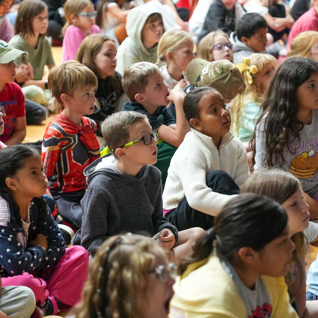 Image 3: A group of elementary school students sitting cross-legged on the floor, attentively looking toward the stage during a school assembly.
