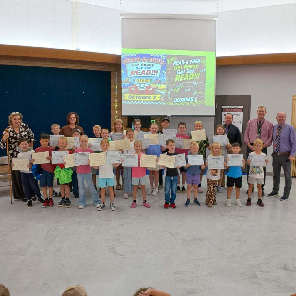 Image 10: A wide group photo of elementary students holding their certificates alongside school staff and administrators standing in front of a screen promoting a school Read-A-Thon event.