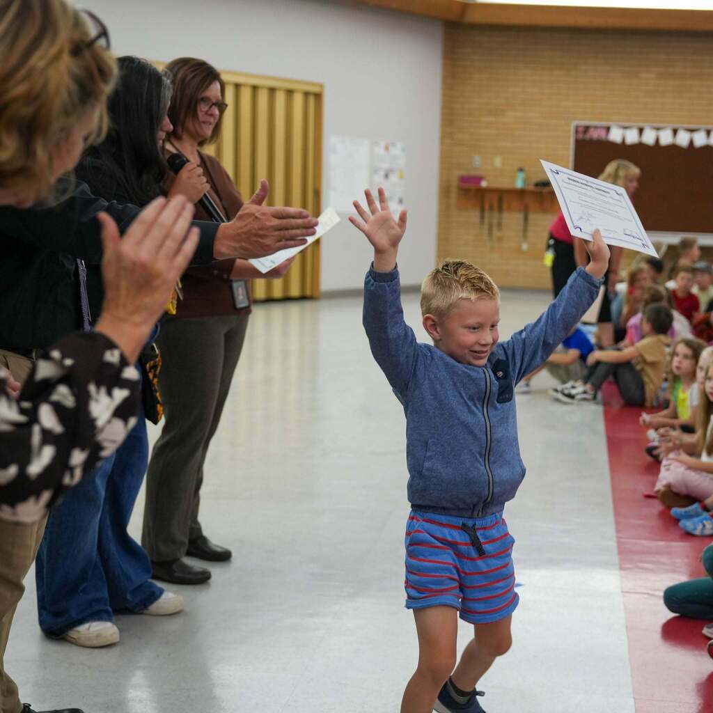 Image 1: A excited young elementary student wearing a blue jacket and striped shorts smiling and raising both arms—holding a certificate in one hand—as school staff clap and cheer for him.