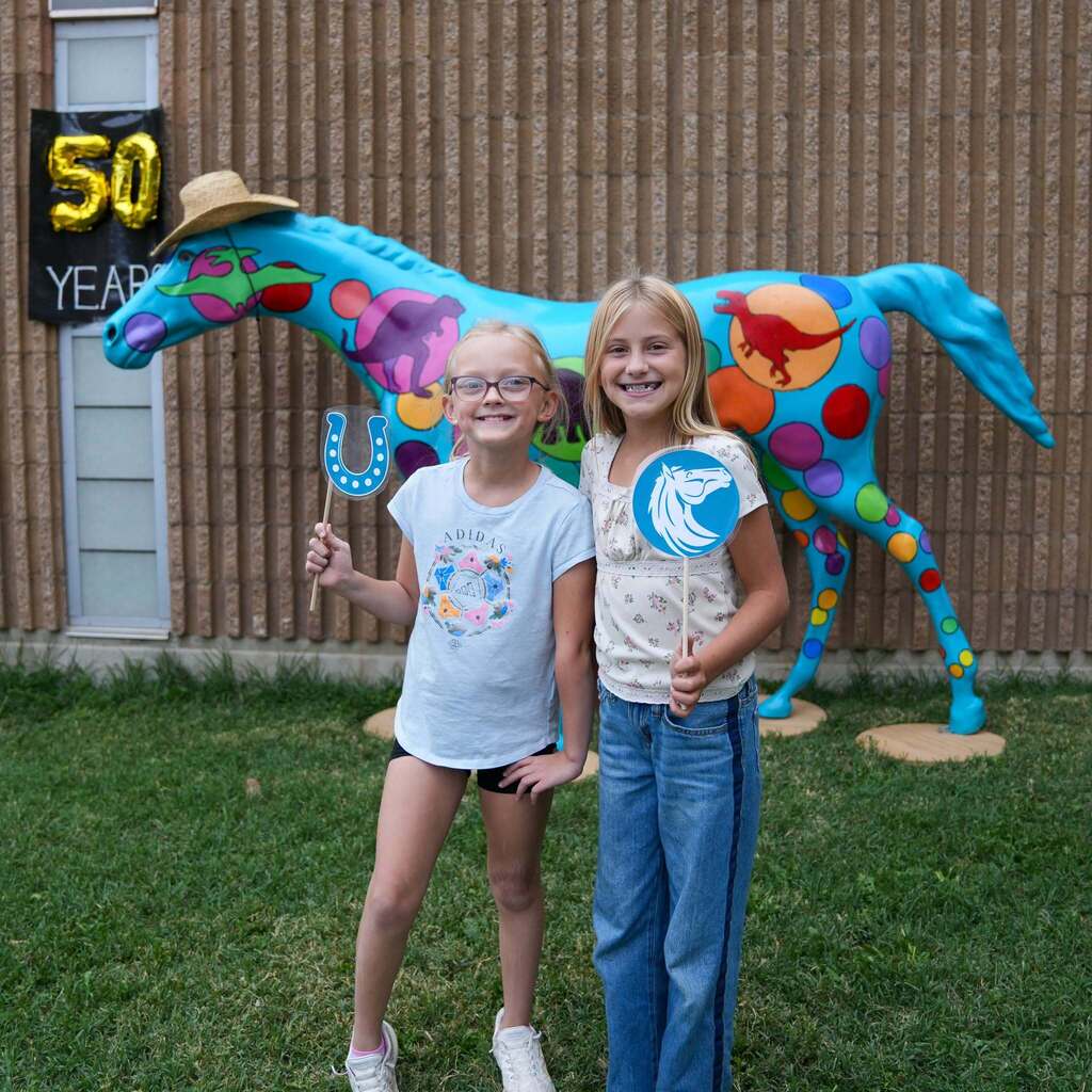 Image 9: Two smiling students holding photo booth props in front of a colorful, painted horse statue celebrating a 50-year school milestone.