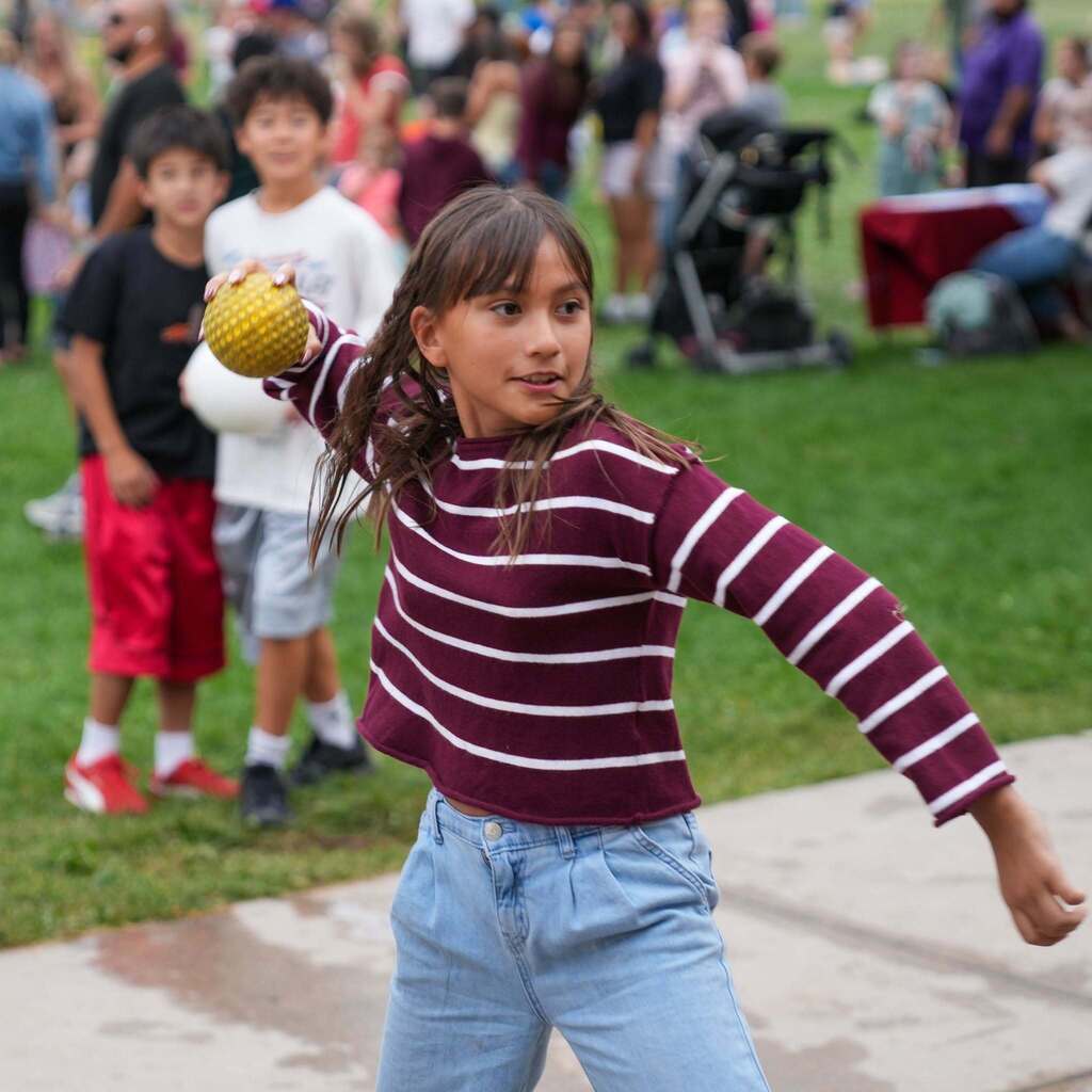 Image 7: A young student wearing a striped shirt aiming and preparing to toss a yellow ball during an outdoor school carnival game.
