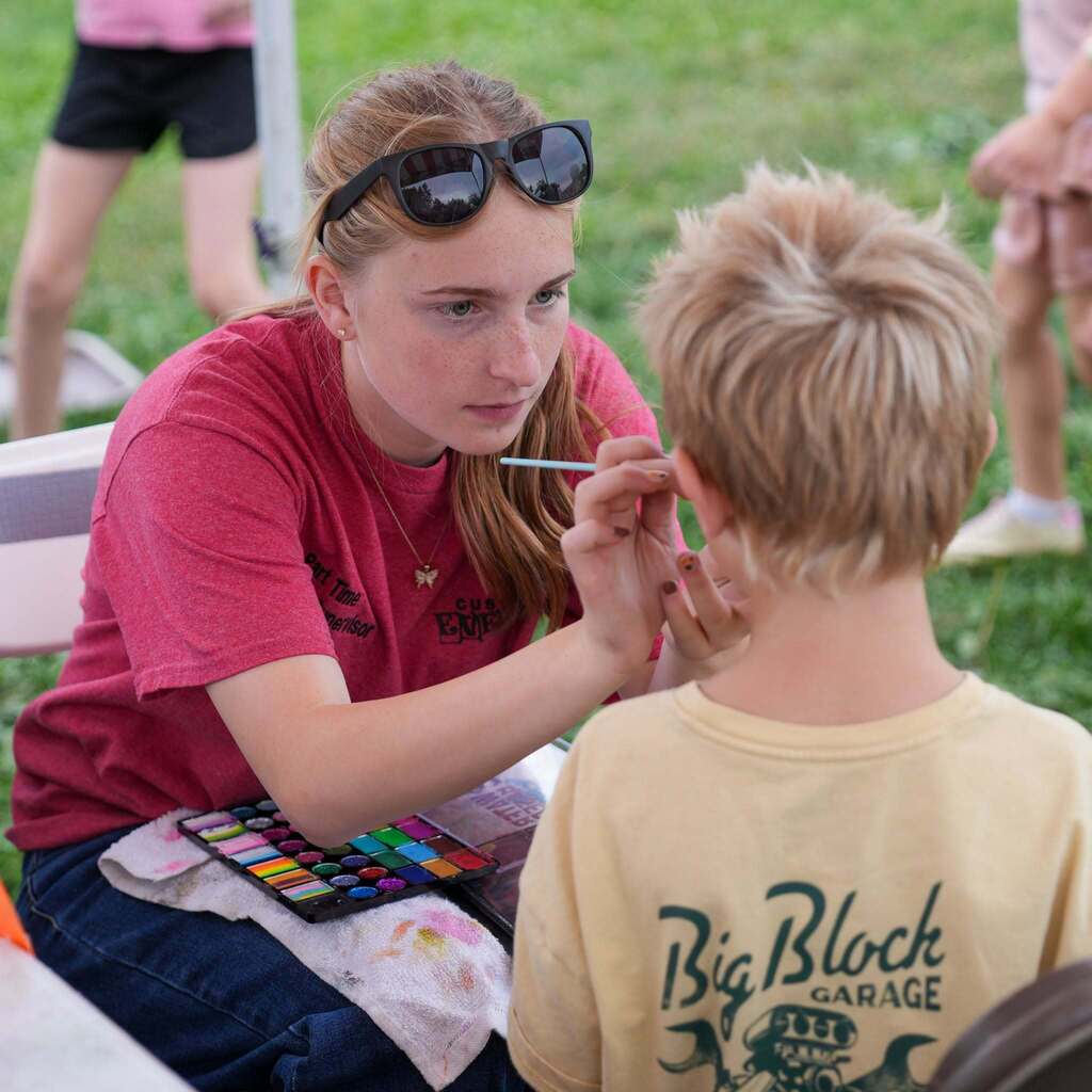 Image 5: A staff member carefully painting a design on a young student's face at an outdoor event booth.