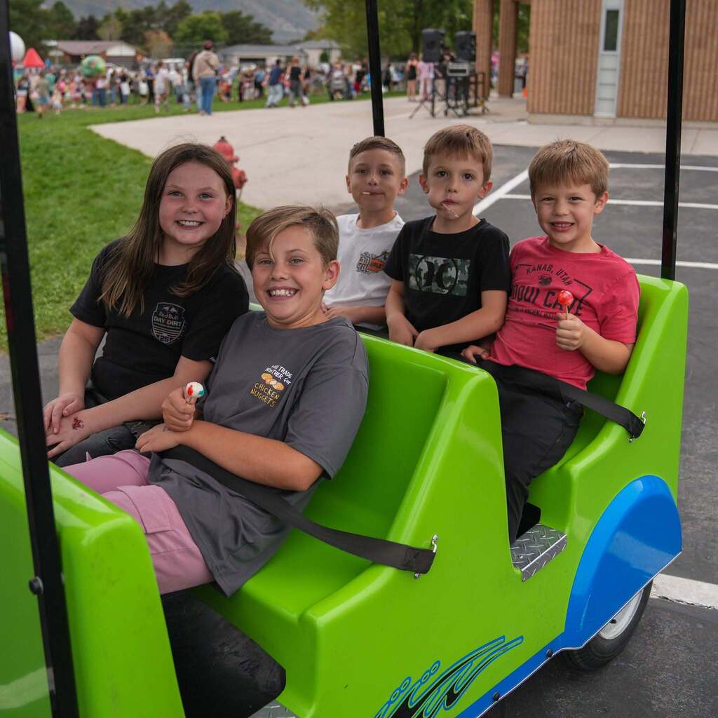 Image 8: Five young students sitting together and smiling inside a bright green event cart while holding lollipops.