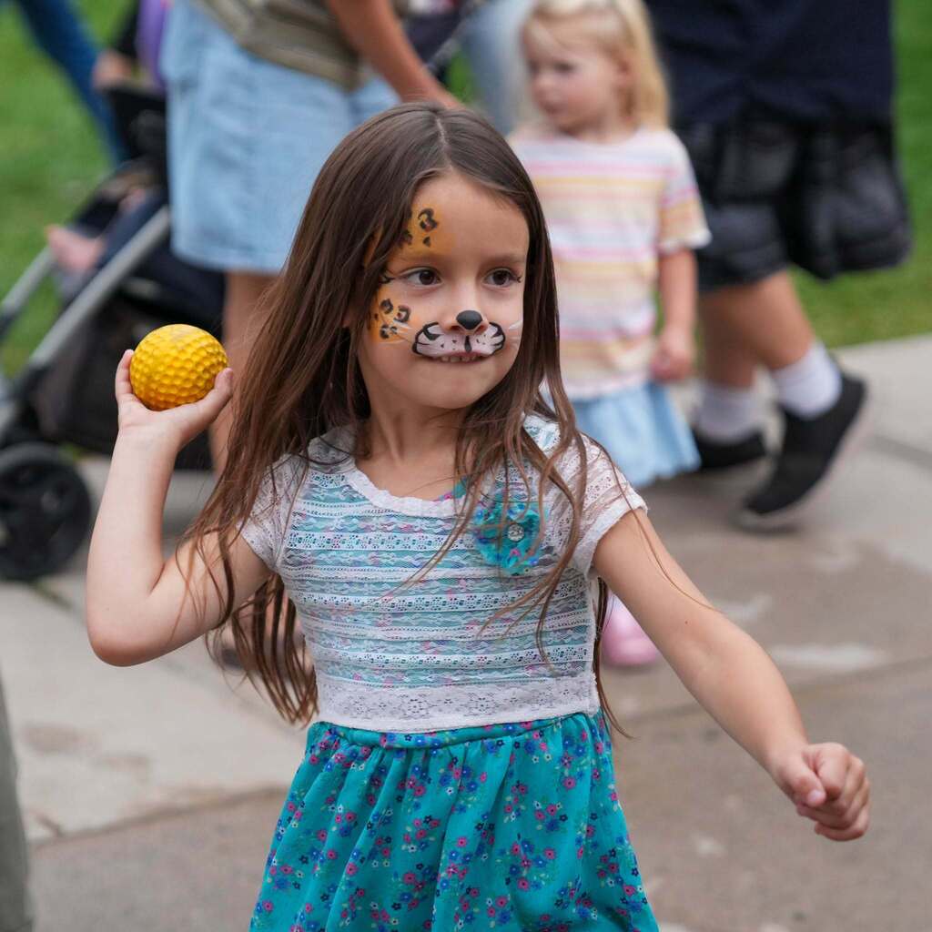 Image 1: A young girl with cheetah face paint smiling and winding up to throw a yellow ball during an outdoor school carnival activity.