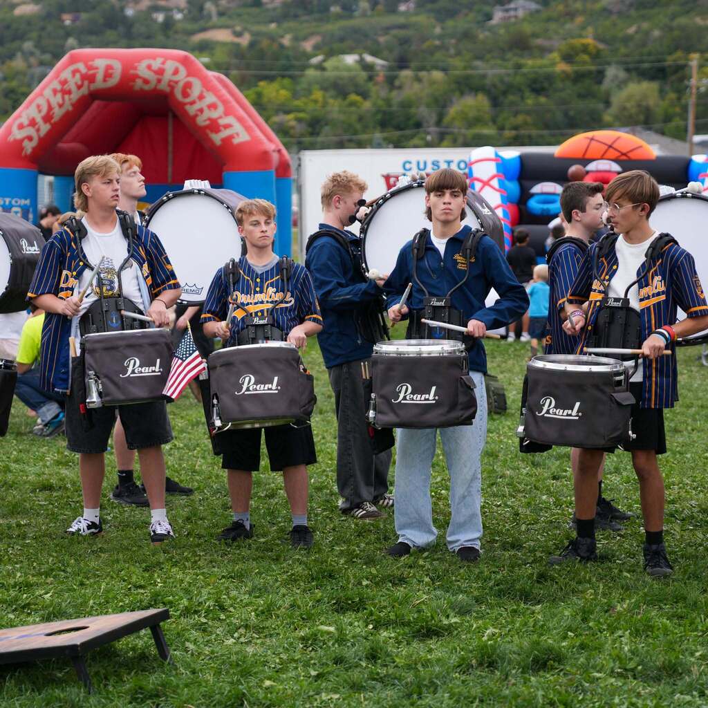 Image 3: Members of a school drumline wearing matching pinstriped baseball-style jerseys and playing snare drums outdoors in front of inflatable event attractions.
