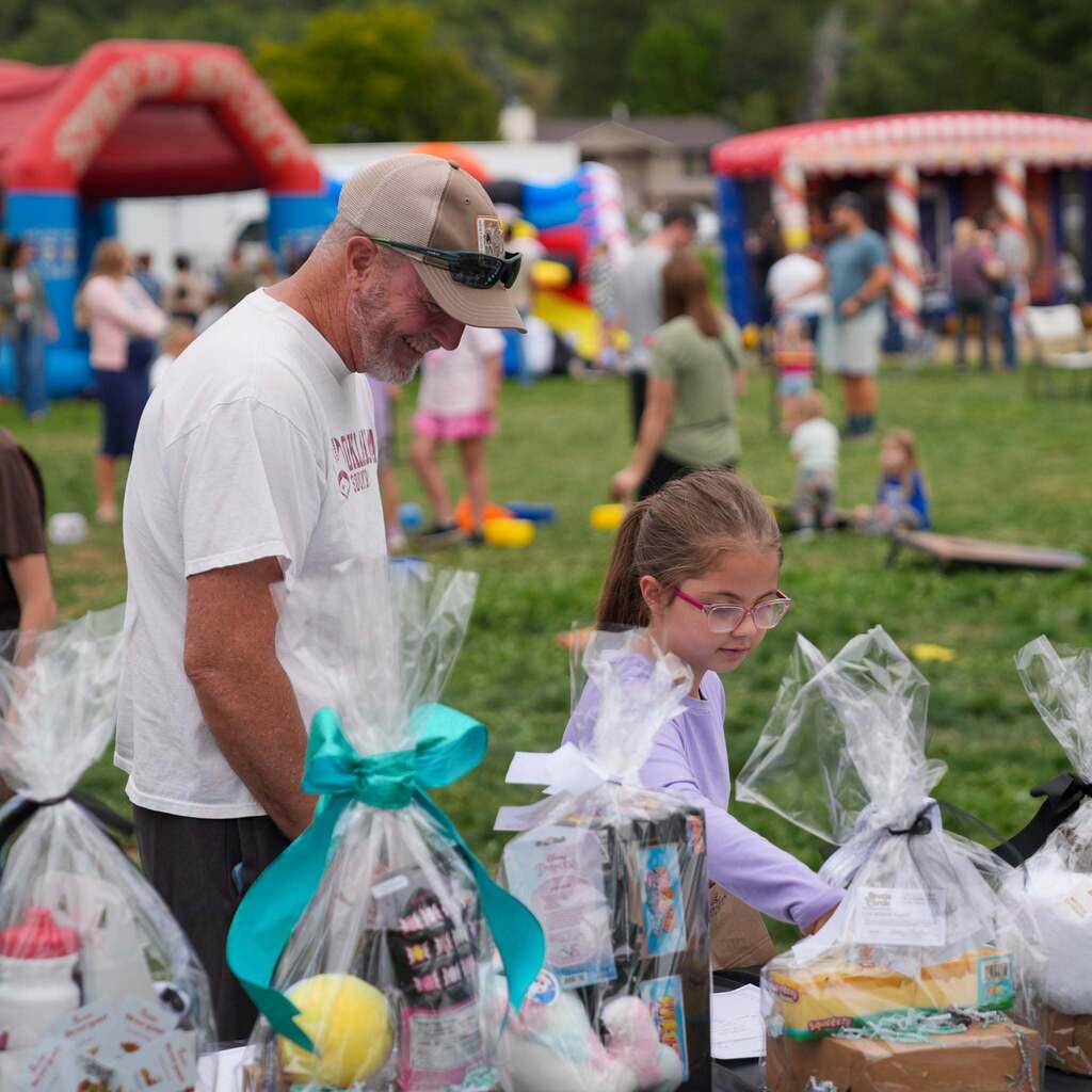 Image 10: A smiling adult and a young student with glasses looking over wrapped gift baskets displayed on a table at an outdoor school event.