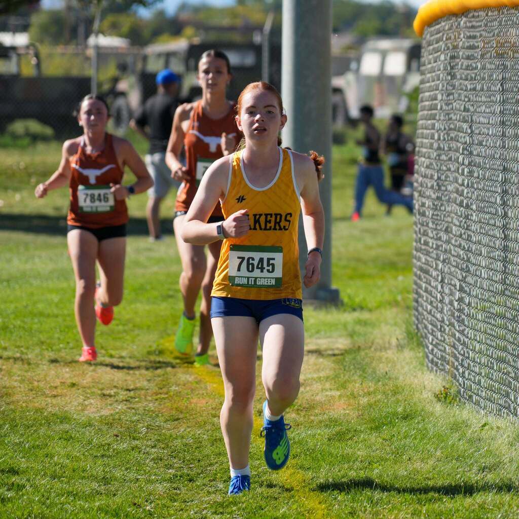 Image 3: A female cross-country runner wearing a yellow and navy "Lakers" jersey running along a grassy course alongside a chain-link fence.
