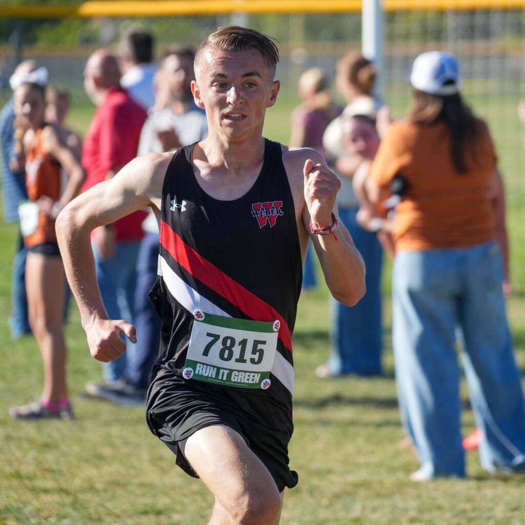Image 7: A male student-athlete in a black and red Weber High uniform sprinting hard toward the finish line during a race.