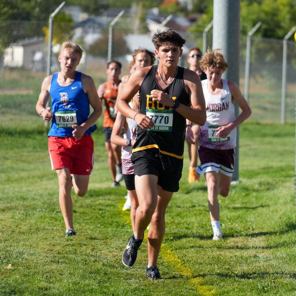 Image 6: A male cross-country runner in a black singlet leading a group of competitors along a marked grass track.