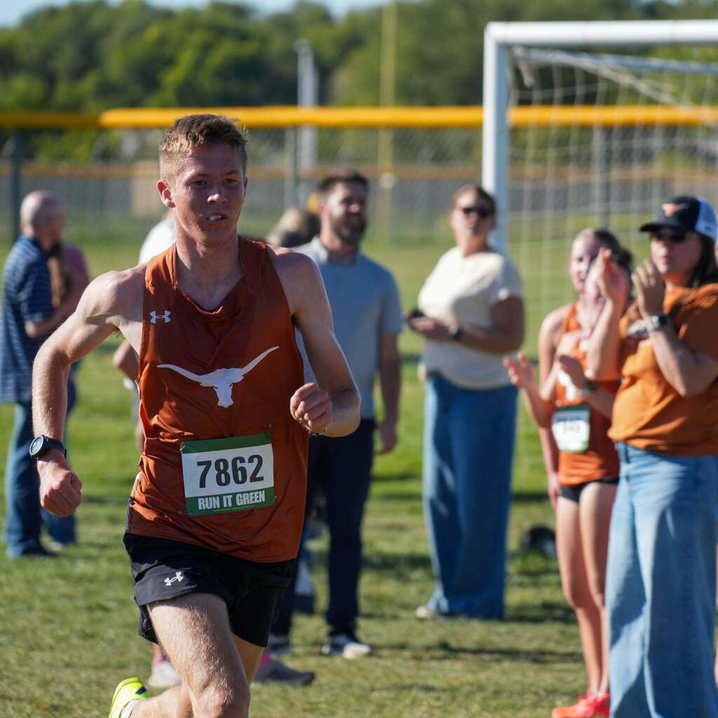 Image 9: A male cross-country runner in a rust-orange jersey pushing through a stride while spectators clap and cheer for him from the sidelines.