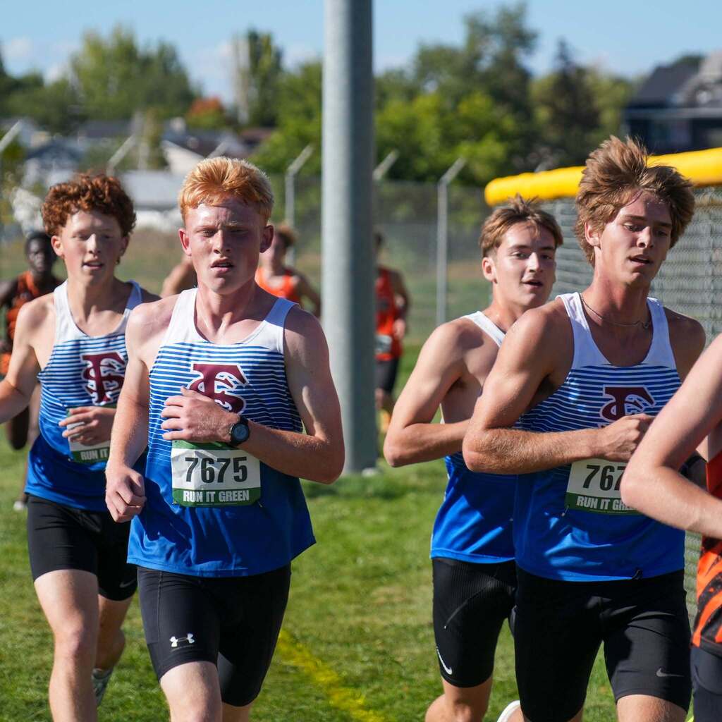 Image 5: A pack of male student-athletes in blue and white striped uniforms running together in a tight group during a cross-country race.