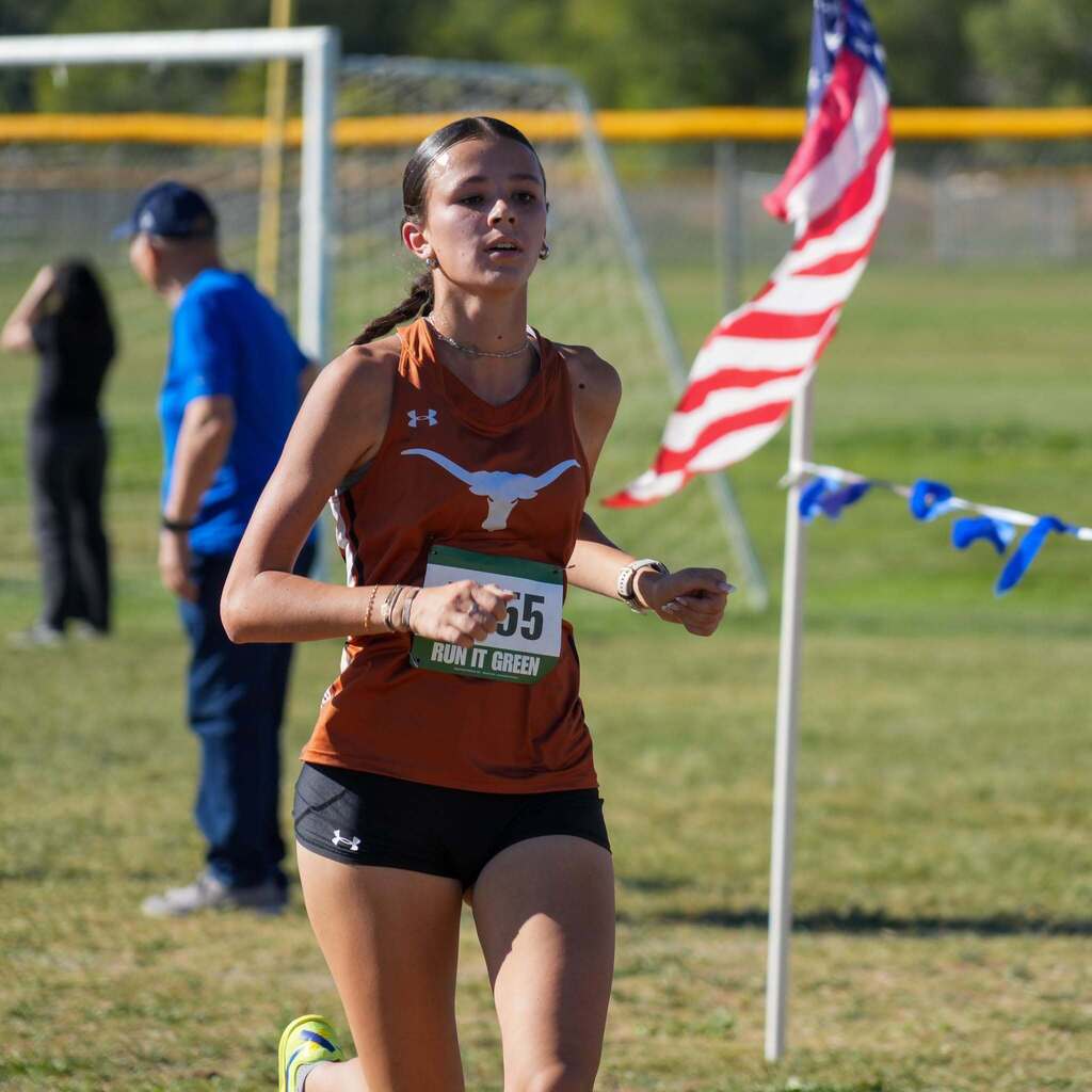 Image 4: A female cross-country athlete in a rust-orange tank top running focused along an outdoor grass course near an American flag.