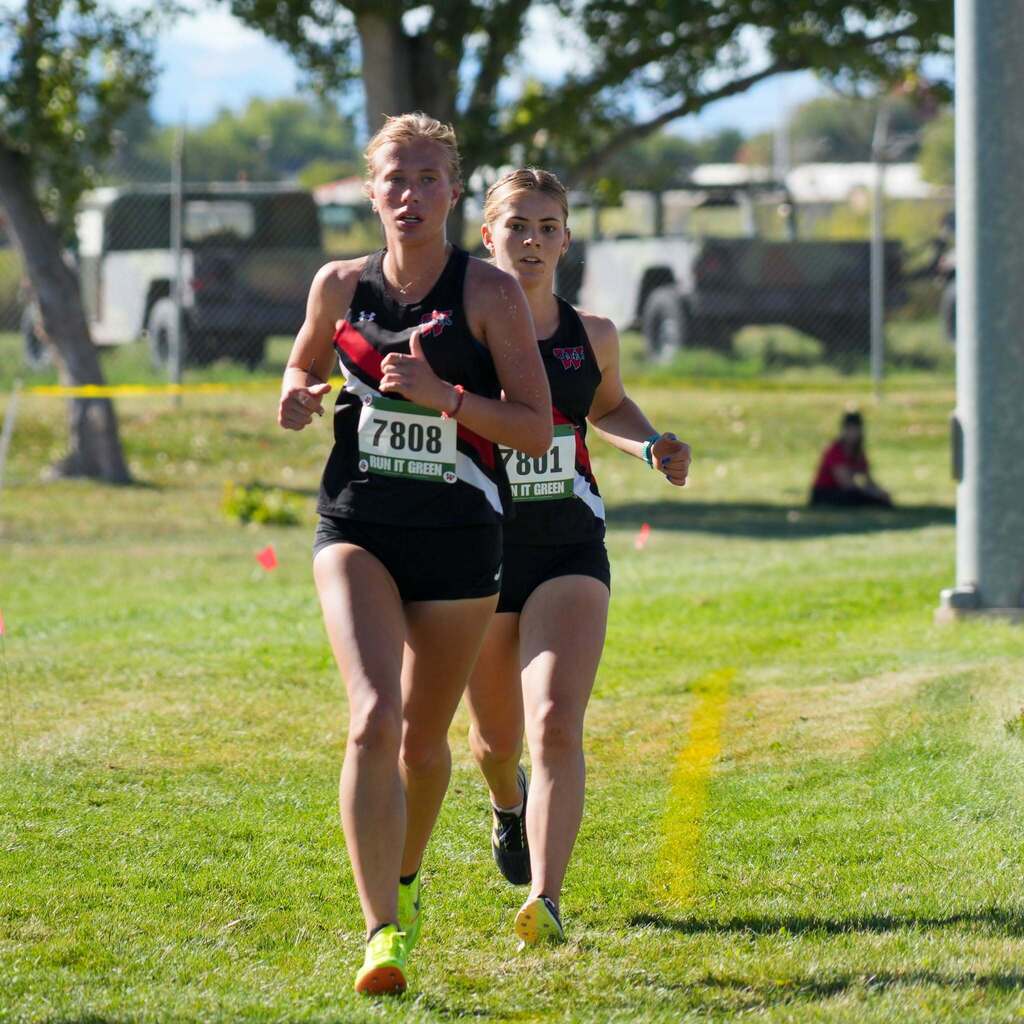 Image 8: Two female Weber High cross-country runners in black singlets pacing together along a sunny, open grass course.