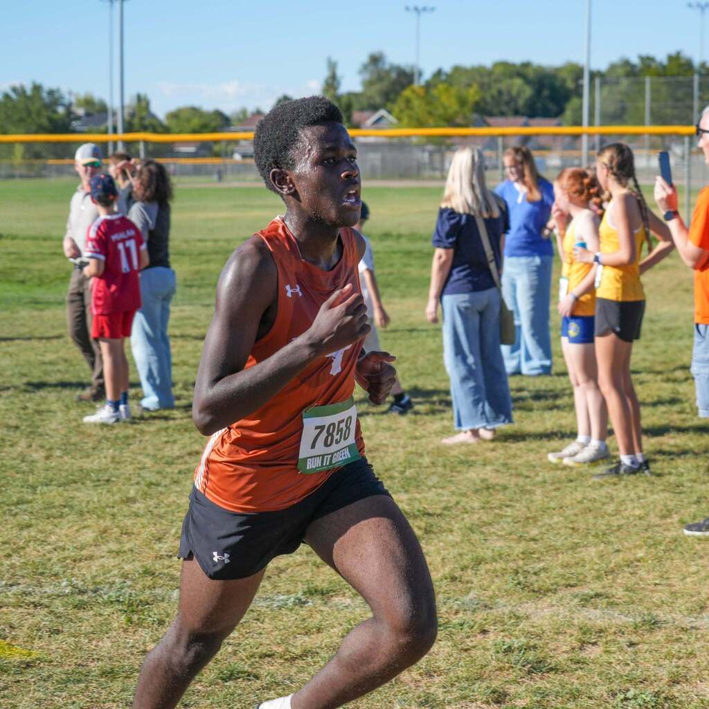 Image 10: A focused male runner in a rust-orange tank top competing on a grassy field with spectators watching in the background.