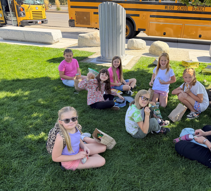 Fourth-graders sitting on the green grass outside enjoying sack lunches with a yellow Weber School District bus in the background.