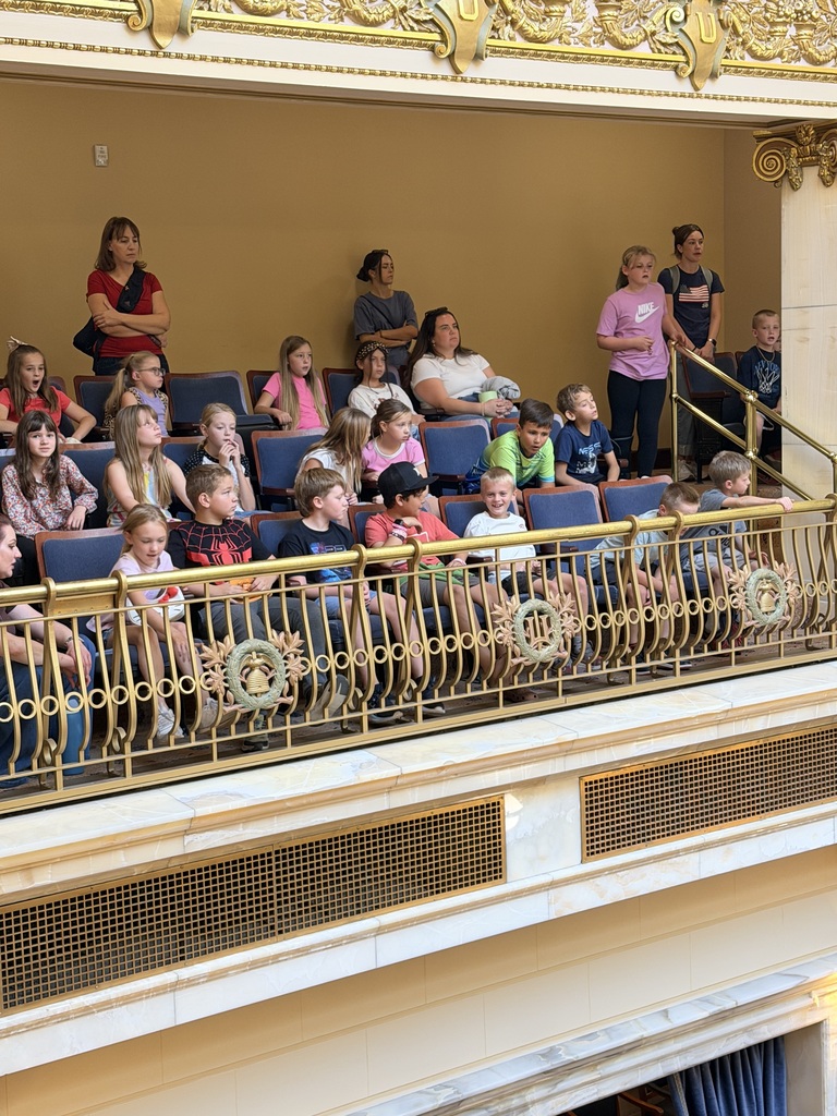 Students seated in balcony rows watching a presentation inside a Capitol legislative chamber.