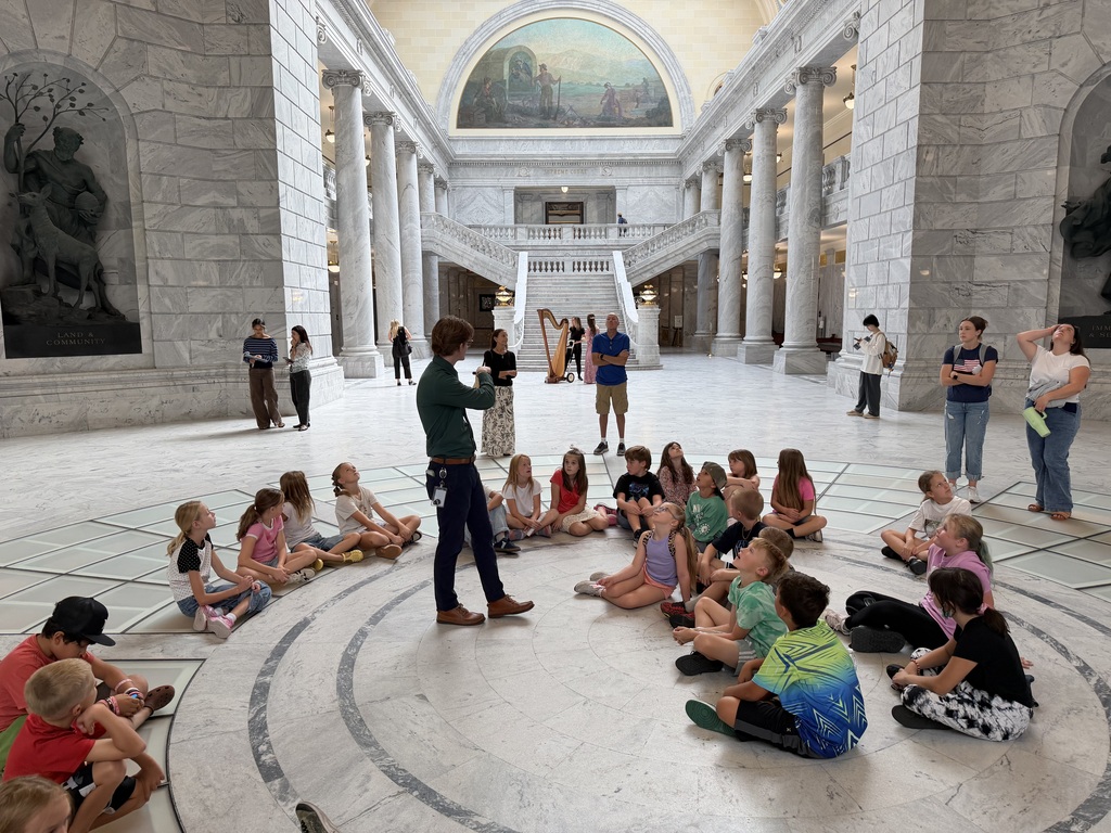 Students seated in a circle on the polished floor of the grand marble rotunda at the Utah State Capitol listening to a tour guide.