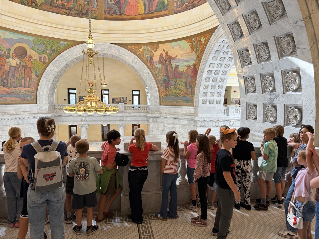 Fourth-graders leaning over a marble balcony railing inside the Capitol to view the dome murals and chandelier above.