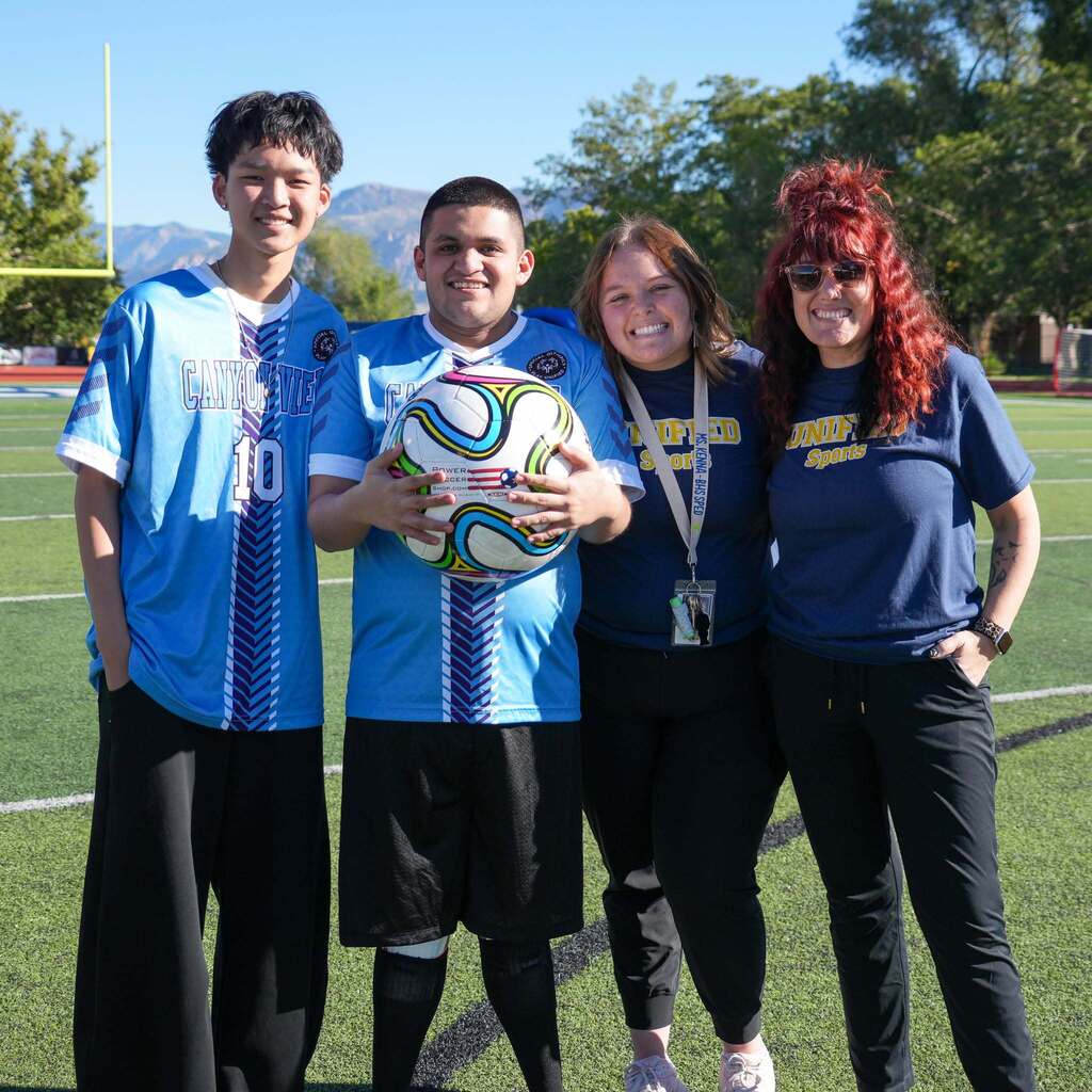 Image 6: Two Canyon View soccer players and two staff members wearing navy Unified Sports shirts smiling together while holding a soccer ball on the field.
