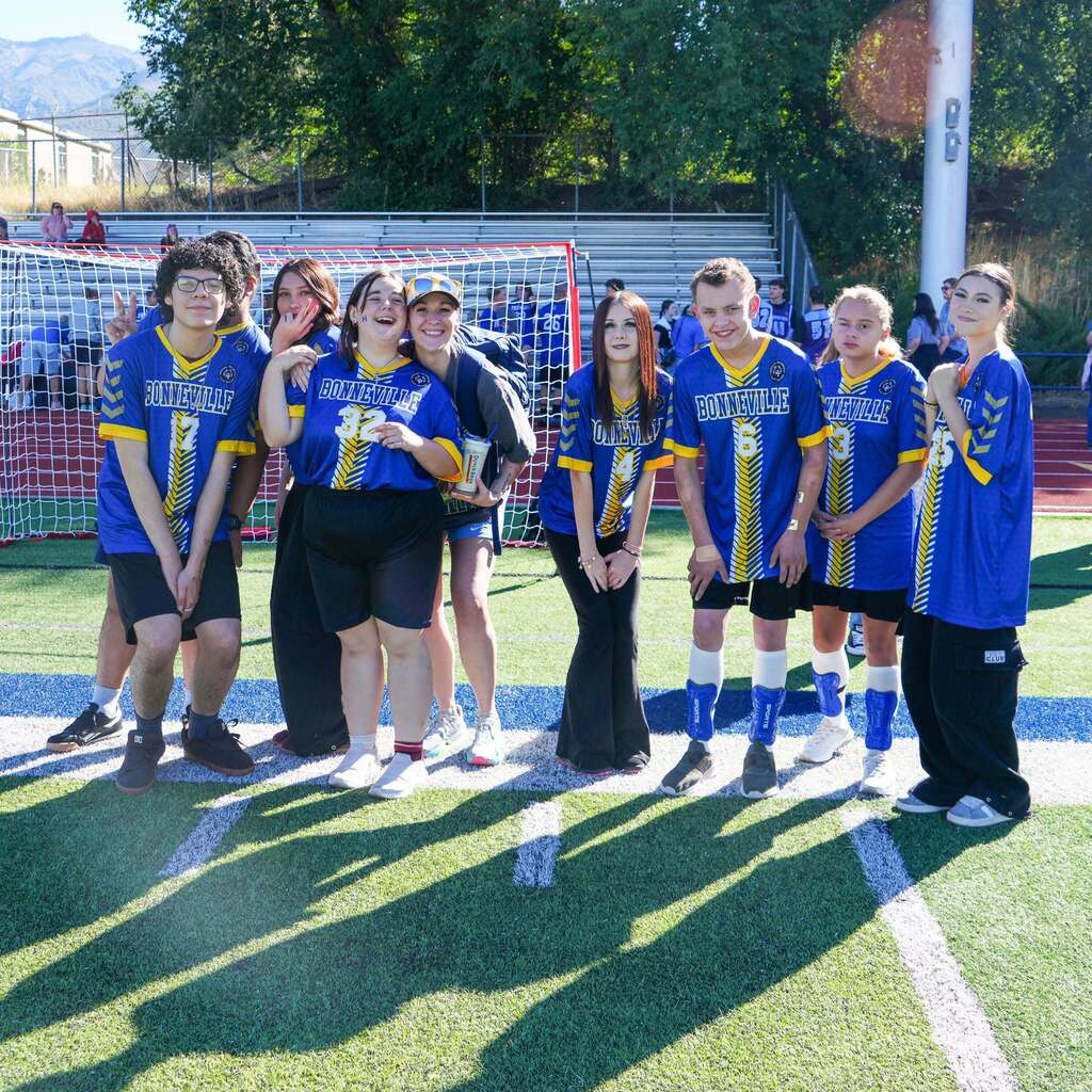 Image 1: A group of Bonneville High Unified Sports athletes and student partners wearing blue and yellow jerseys posing together on the football field gridiron.