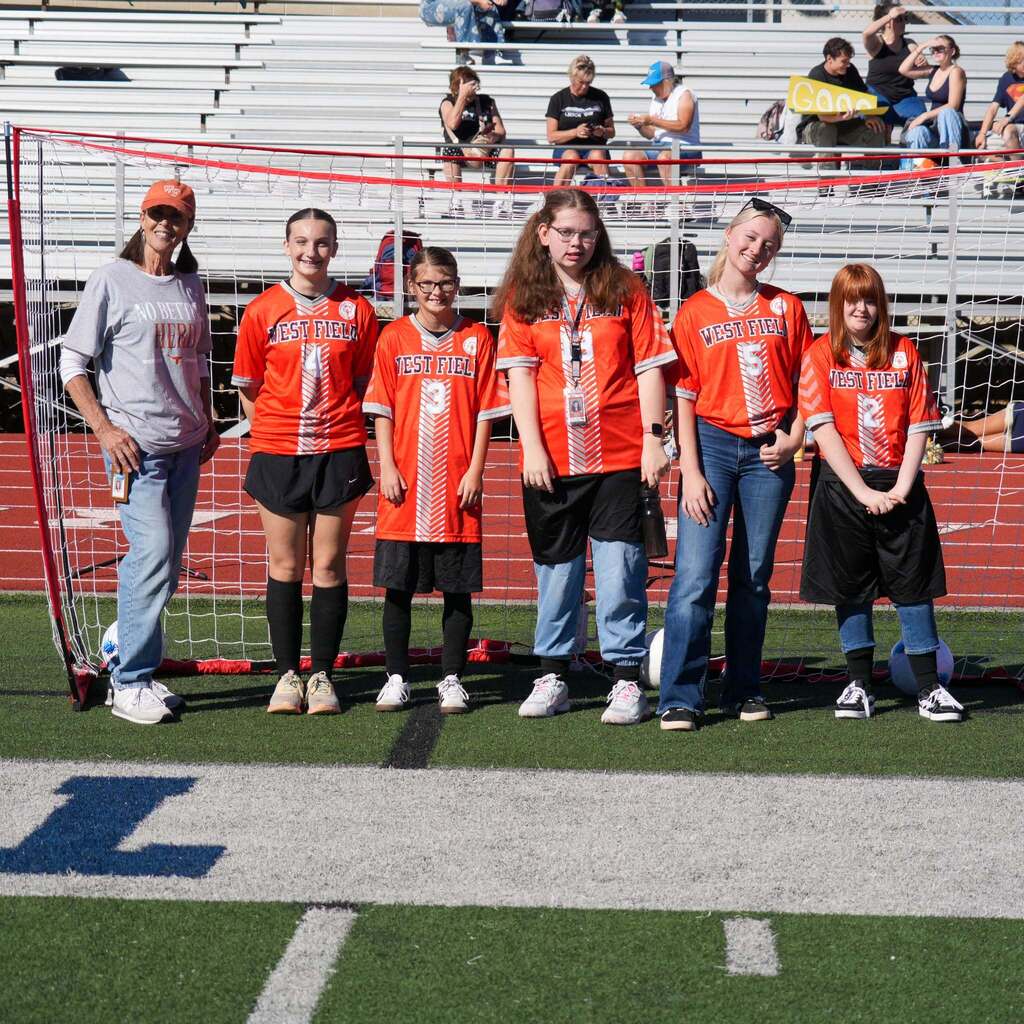 Image 10: A coach and five Westfield High Unified soccer athletes wearing bright orange jerseys standing together in front of a soccer goal net.